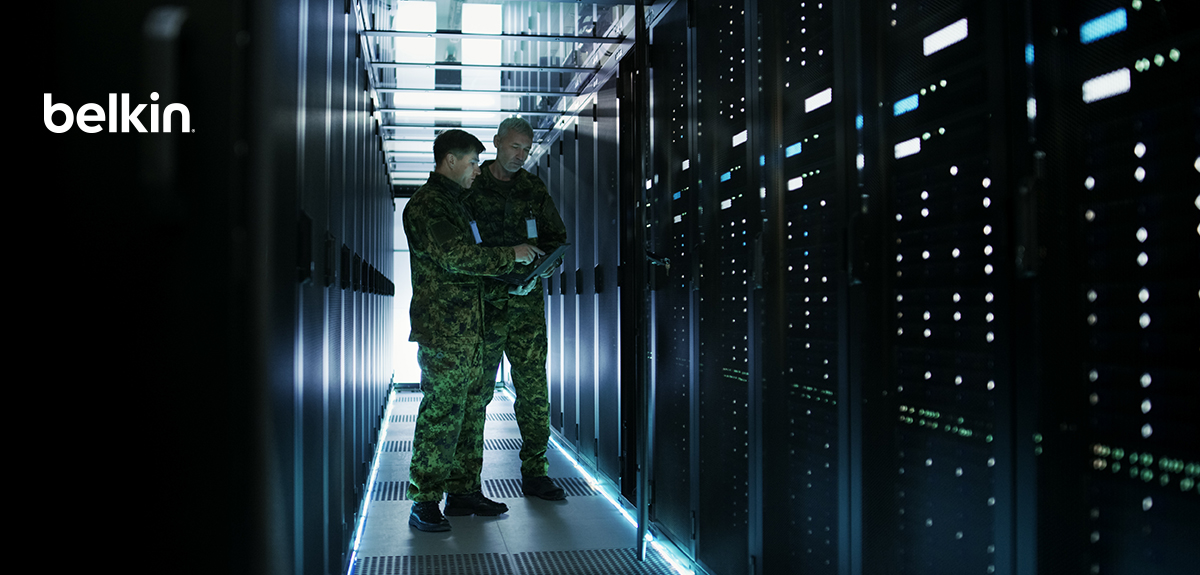 Two military personnel in camouflage uniforms examine a tablet while standing in a high-security data center server room. Rows of illuminated server racks line both sides. The Belkin logo appears in the upper left corner.