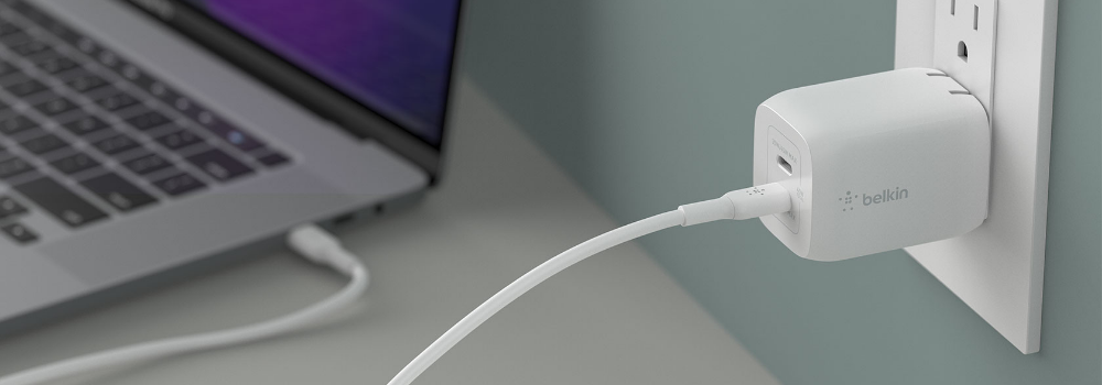 The image shows a white Belkin wall charger plugged into an electrical outlet. A USB-C cable is connected to the charger and extends toward a nearby laptop, which appears to be in use. The setup highlights a clean and organized charging solution, showcasing the charger’s compact design and compatibility with modern devices. The background features a muted green wall and a minimalist workspace.