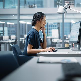 A person seated at a desk in a modern office, focused on a computer screen while working, with a desk lamp and papers nearby.