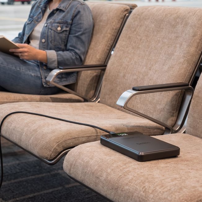 A Belkin power bank showing 100% sits on an airport gate seat while a traveler in a denim jacket reads a book two seats over