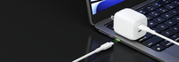 The image shows a close-up of a laptop keyboard with a USB-C cable connected to it. The cable is plugged into a white wall charger with a compact design. The charger is positioned on a reflective black surface, emphasizing its sleek design and compatibility with the laptop. The cable also features a green-lit connector tip, suggesting a functional or aesthetic design element.