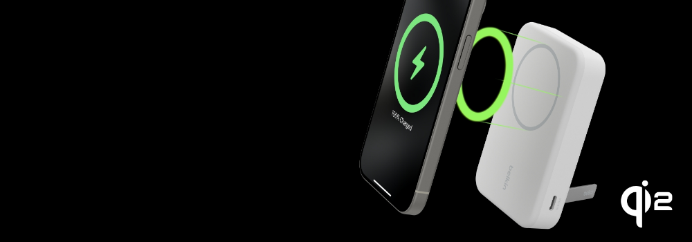 A Belkin Qi2 wireless charger with a magnetic attachment is shown charging a smartphone. The phone's screen displays a green charging icon with '100% Charged.' The charger is white with a foldable stand and a USB-C port. The Qi2 logo is visible in the bottom right corner on a black background.