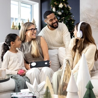 A family of four sits together on a couch in a cozy, holiday-decorated room, smiling and sharing tech gadgets.