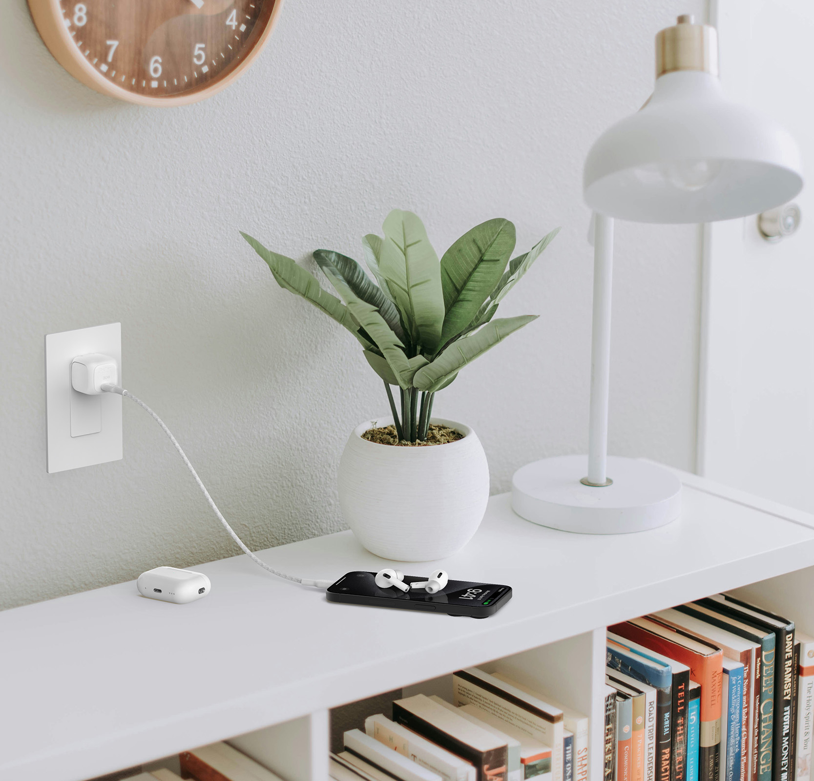 The image shows the Belkin BoostCharge Compact USB-C Wall Charger 30W plugged into a wall outlet, fast charging an iPhone on a nearby table with AirPods placed on top, alongside a small potted plant.