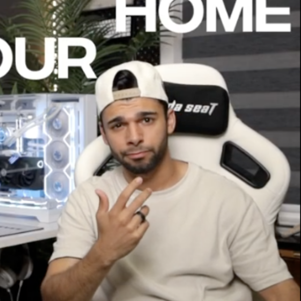 Jason Hymanson in a light beige t-shirt and backwards cap sits in a gaming chair, gesturing with his hand in a modern home office setup featuring a glowing custom-built PC, dual monitors, and providing product reviews for Belkin.