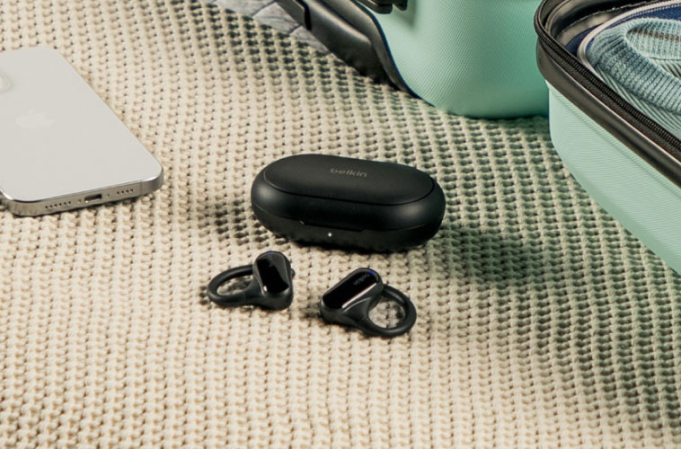 Pair of wireless Belkin earbuds with their charging case placed next to a smartphone and open mint green suitcase on a textured beige surface.