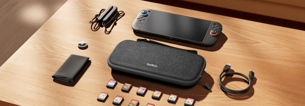A neatly arranged gaming setup on a wooden table featuring a handheld gaming console, a Belkin-branded carrying case, multiple game cartridges, a USB charging cable, a pair of detachable controllers with straps, a compact game card holder, and a small round accessory. The setup highlights organization and portability for gaming on the go.