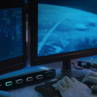 A person types on a keyboard in front of multiple computer monitors displaying cybersecurity and satellite tracking interfaces, with a focus on a device featuring multiple input/output ports below the screens.