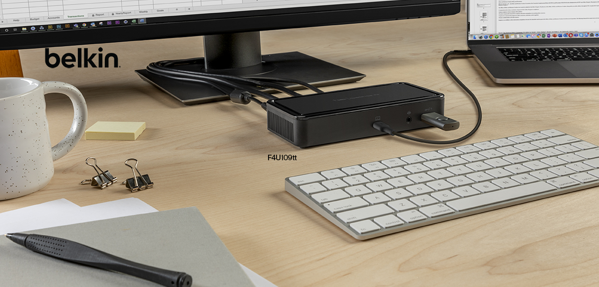 This image features a modern desk setup with a Belkin docking station connected to a laptop and a monitor. The docking station is sleek and compact, with multiple connected cables and a USB device plugged in, emphasizing its versatility and functionality. The desk includes a white keyboard, a speckled coffee mug, sticky notes, binder clips, and a pen on a notebook, creating an organized and professional workspace. The Belkin logo is visible on the monitor's left side.