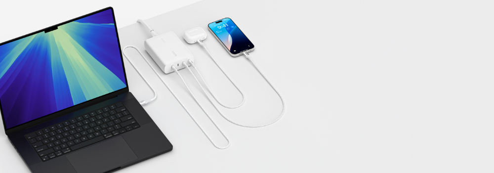 The image shows a workspace setup with a laptop, a multi-port charging hub, and a smartphone. The laptop, with a vibrant blue and green display, is connected to the charging hub via a cable. The white charging hub is placed on a clean, minimalistic desk and powers multiple devices simultaneously, including the smartphone on the right, which displays the lock screen with the time '9:41.' The cables are neatly organized, emphasizing a tidy and efficient charging solution. The background is white, reinforcing a clean and modern aesthetic.
