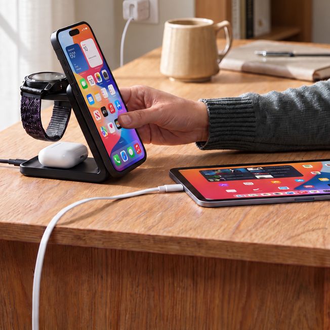 A person reaches for a phone on a Belkin 3-in-1 magnetic charging stand that also charges a watch and earbuds, beside a tablet charging from a white USB-C cable on a wooden desk