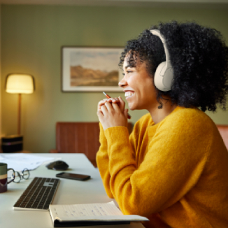 The image depicts a smiling individual with curly hair, wearing headphones, and sitting at a desk with a keyboard, notebook, and pen. The background features a lamp and framed artwork, creating a warm and cozy workspace.