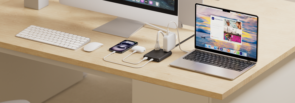 A tidy desk setup featuring a desktop computer, laptop, keyboard, mouse, and smartphone, all powered through a compact multi-port charging hub placed between the devices. Several cables connect the hub to the laptop, phone, and other electronics on the light wood surface.