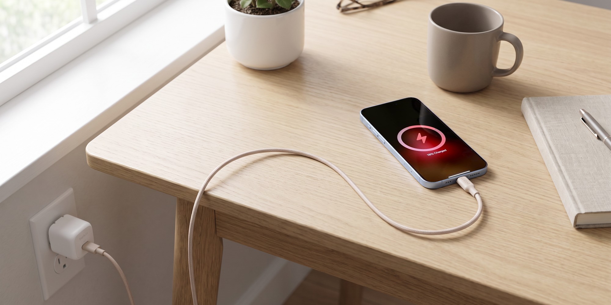 A white Belkin compact USB-C wall charger plugged into a wall outlet, its braided cable running across a light oak desk to an iPhone lying face up with its charging screen on, next to a small plant, a mug and a notebook