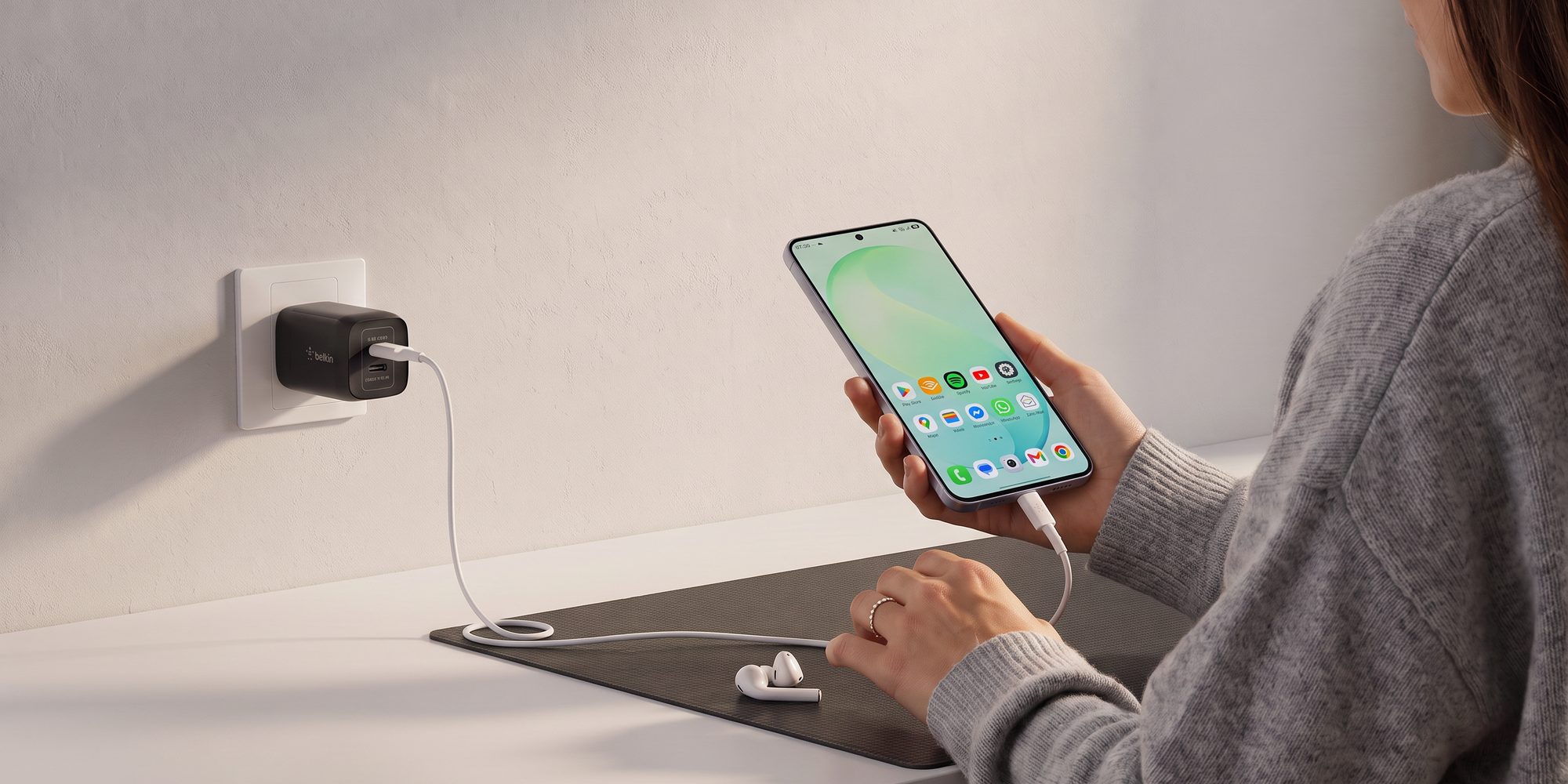 A person holding a phone that is charging from a Belkin compact USB-C wall charger plugged into the wall, with wireless earbuds on the desk beside them