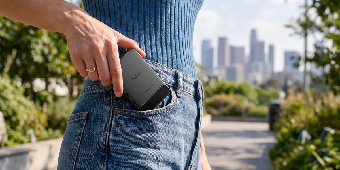 A hand slipping a slim black Belkin power bank into the front pocket of a pair of jeans, with a city skyline out of focus behind