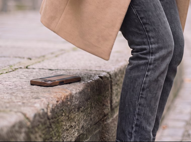 A phone in a case lying on a stone curb behind someone in a camel coat who is walking away, unaware it has slipped out of a pocket